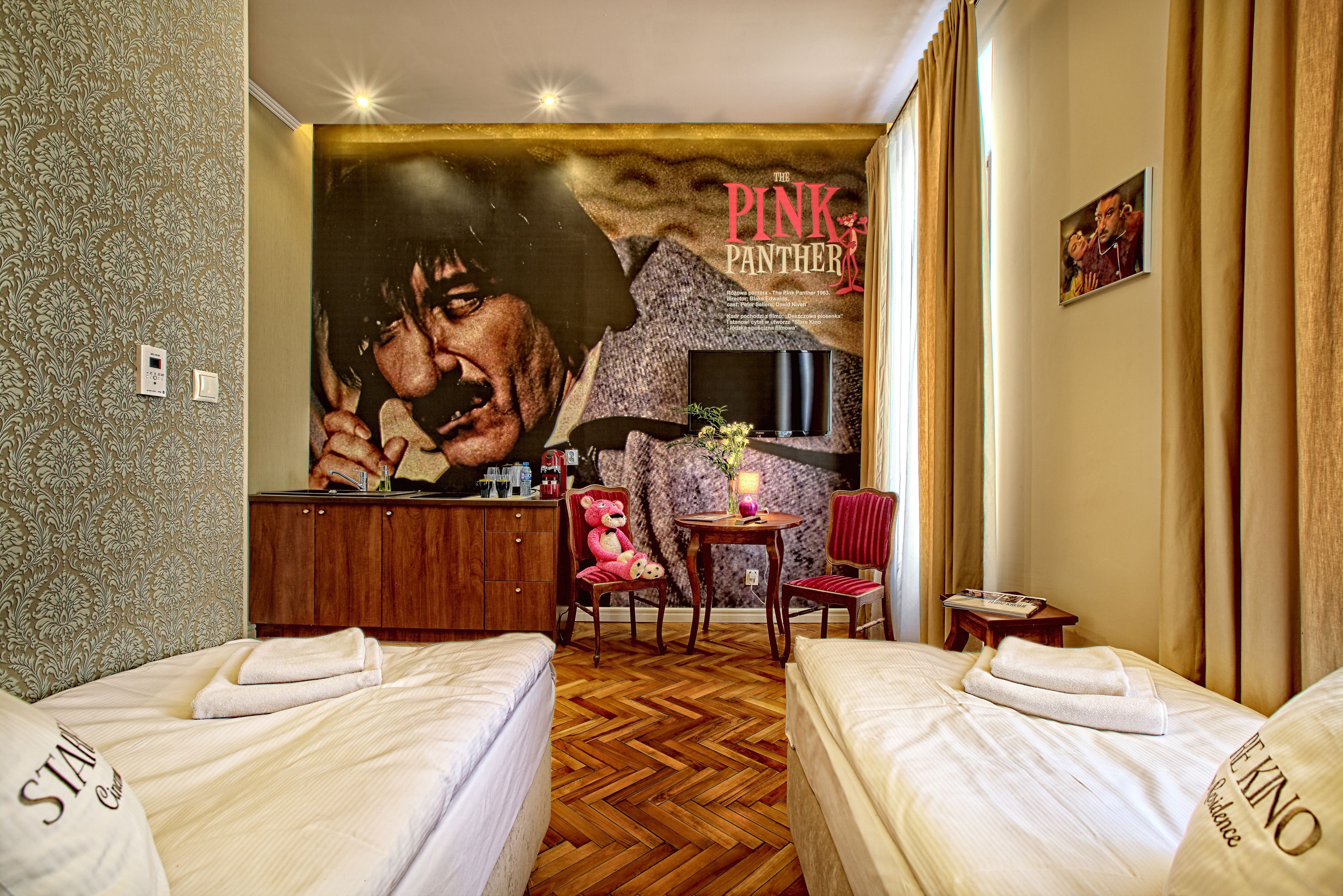A hotel room with two beds, a mural of a man and a woman, a wooden dresser, a small table with a lamp, and a red chair.