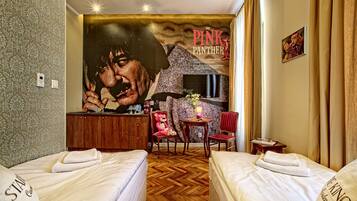 A hotel room with two beds, a mural of a man and a woman, a wooden dresser, a small table with a lamp, and a red chair.