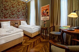 Habitación de hotel con dos camas, piso de madera con diseño espiga, una mesita con lámpara, una silla y un cartel en la pared.