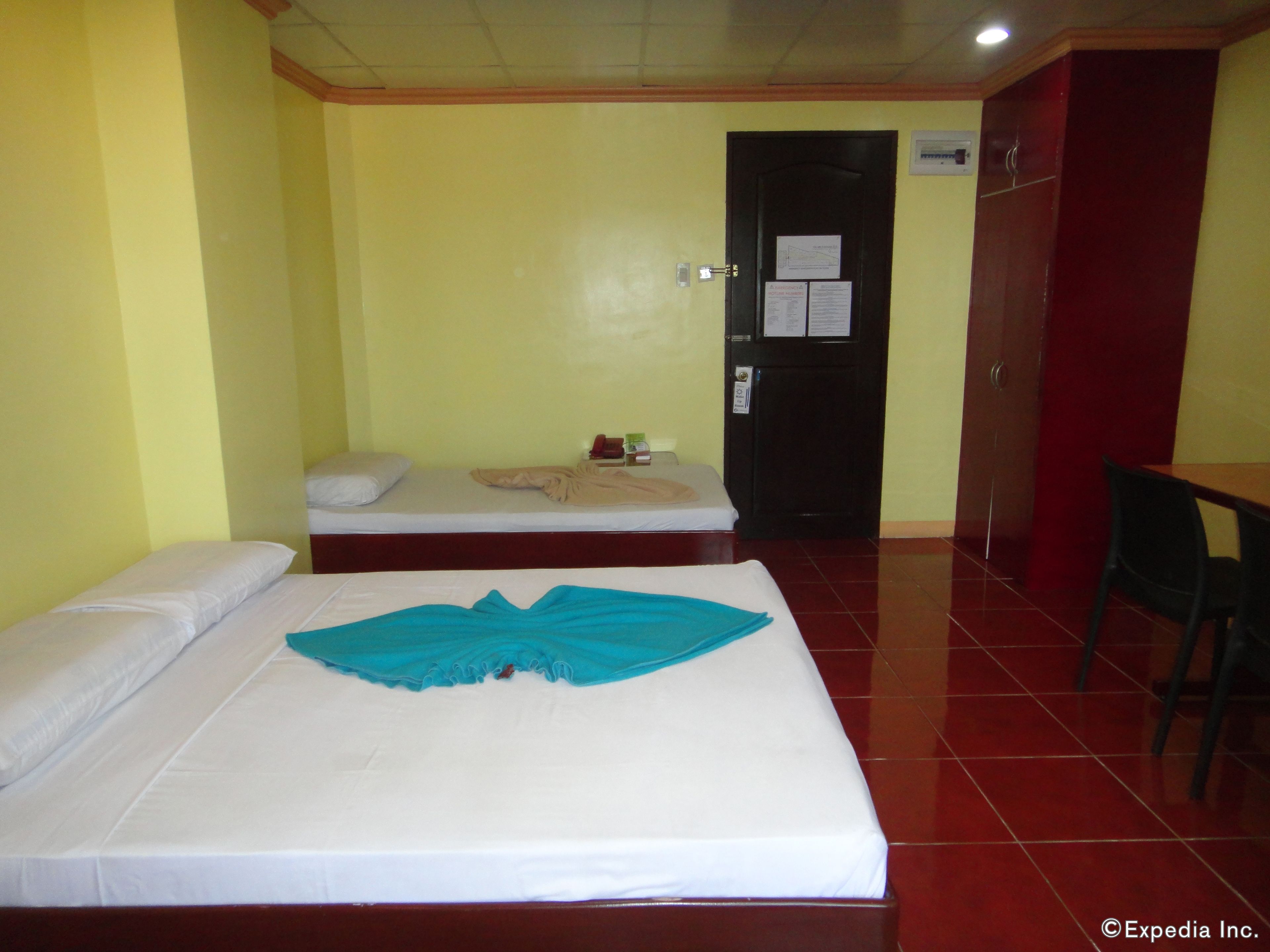 Habitación de hotel con dos camas, una con una manta azul y la otra con sábanas blancas. Hay un escritorio con una silla y una puerta con un aviso.