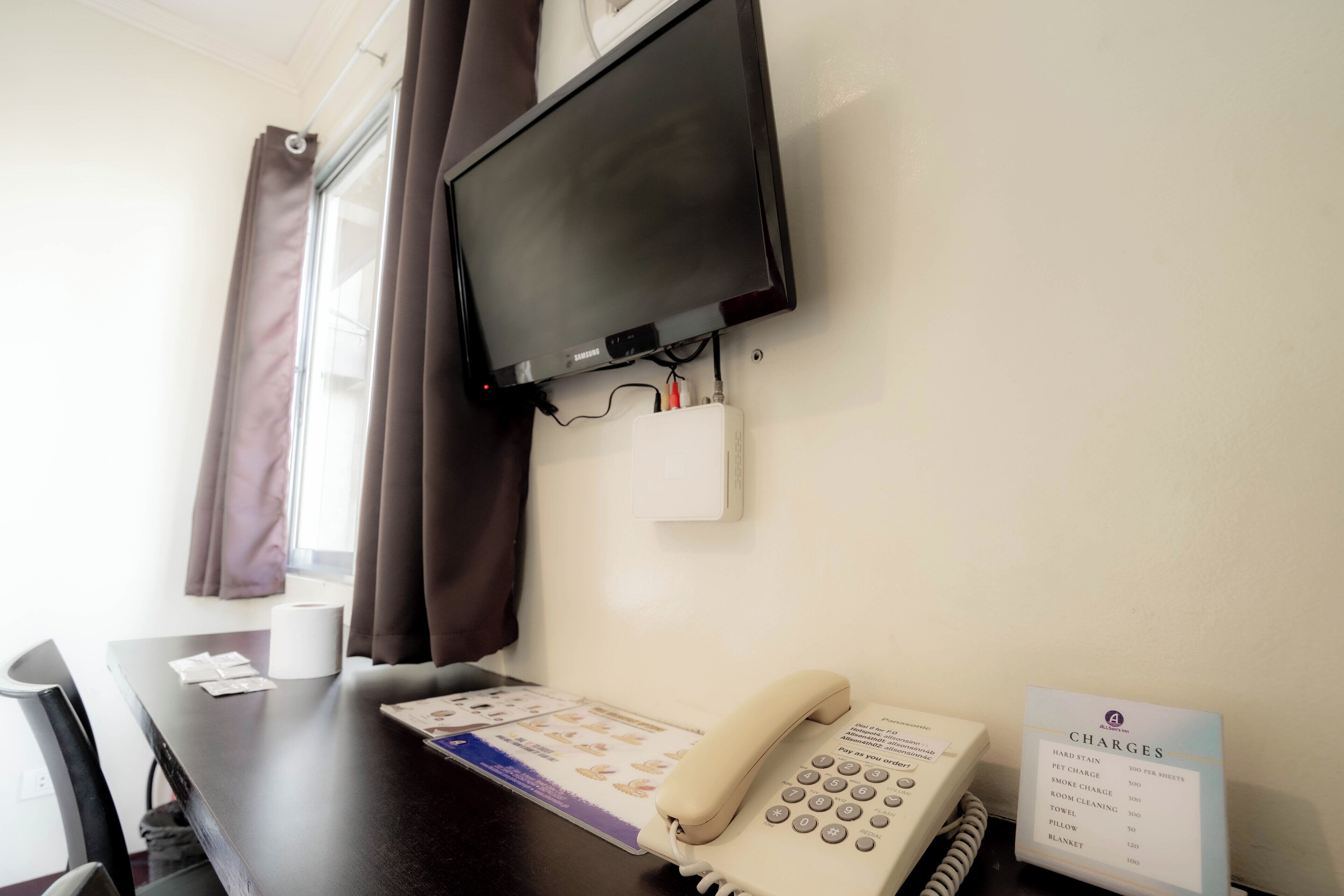 A hotel room with a flat-screen TV mounted on the wall, a telephone, and a sign listing room rates.