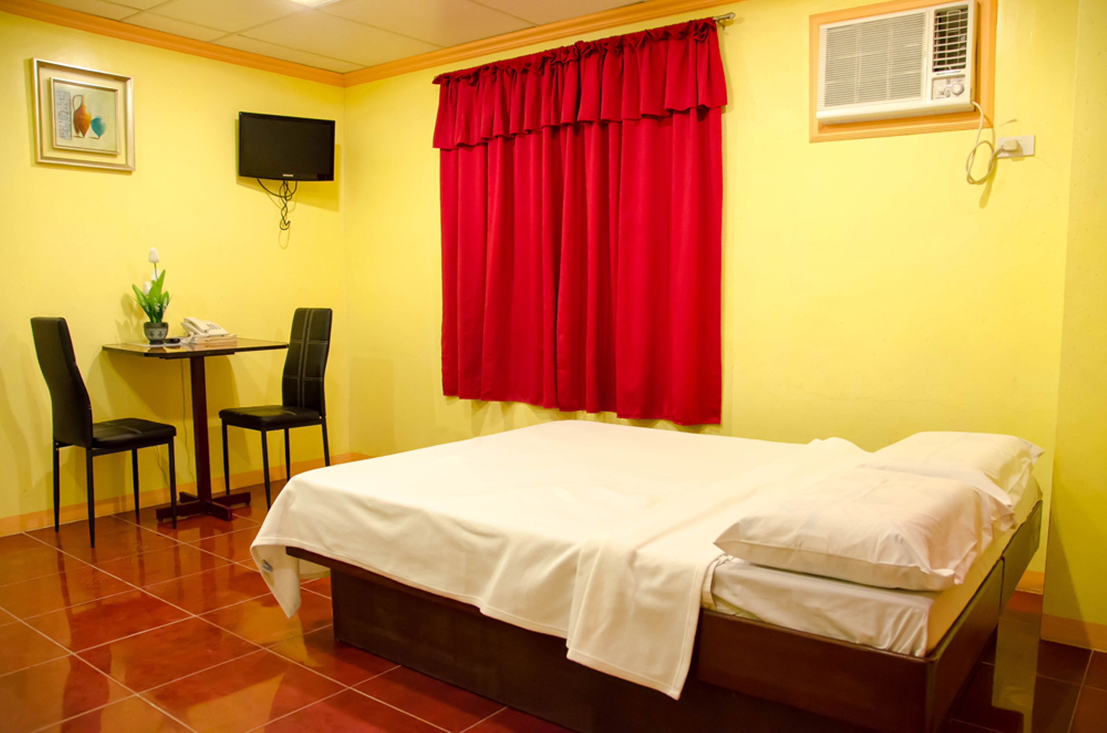 Une chambre d’hôtel avec un lit, deux chaises, une petite table, un téléviseur et une fenêtre avec des rideaux rouges.