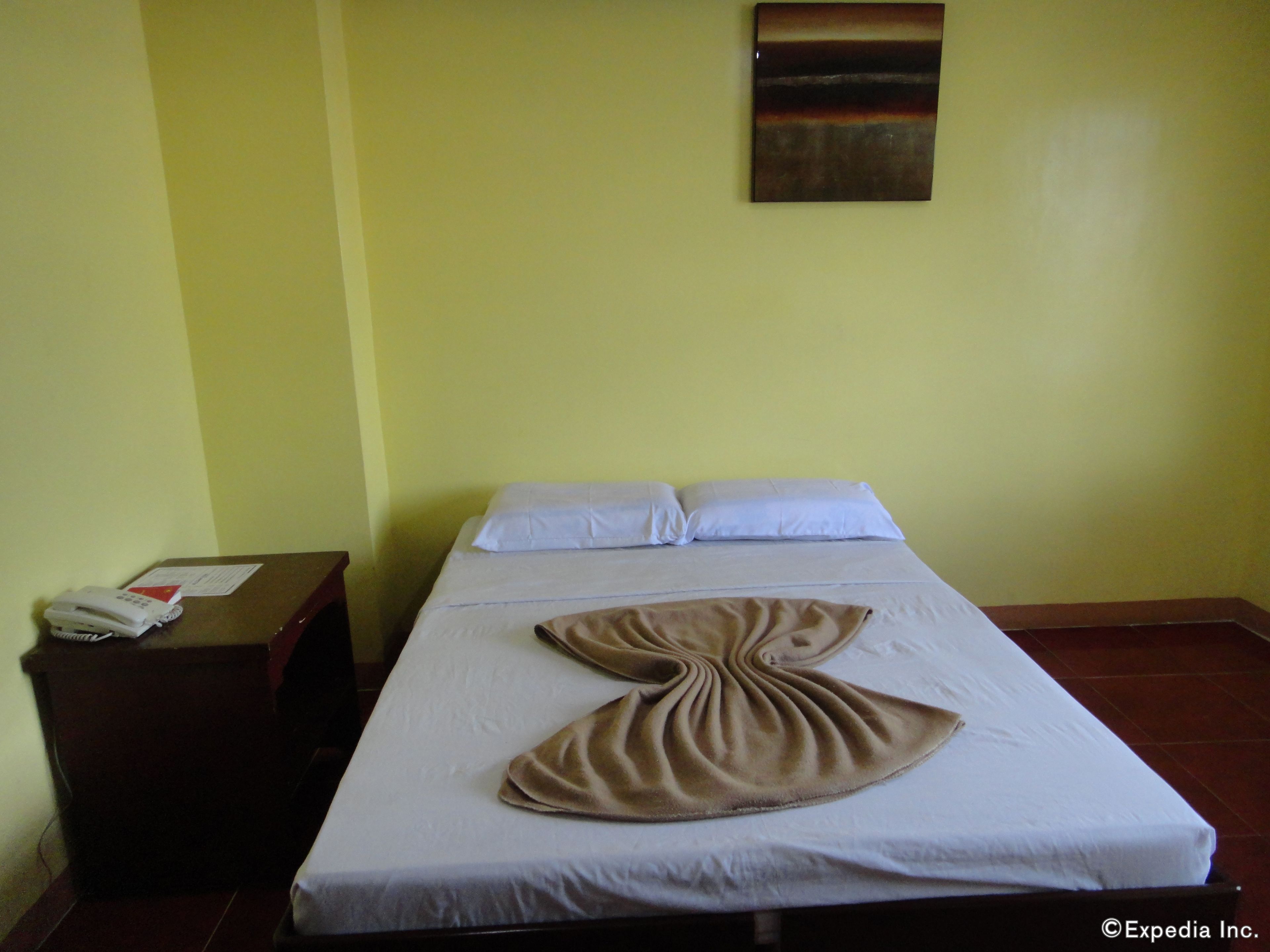 A single bed with white linens and a brown blanket, a bedside table with a phone and papers, and a painting on the wall.