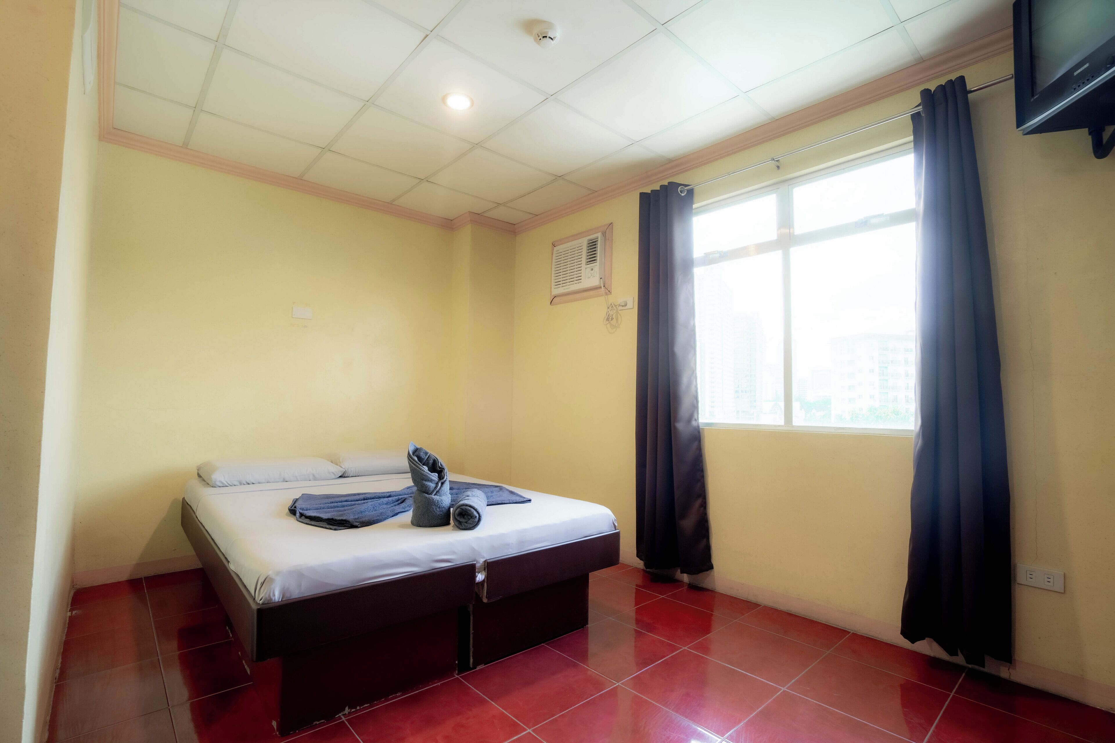 A hotel room with a bed, a window with curtains, a ceiling with recessed lighting, and a wall-mounted TV.