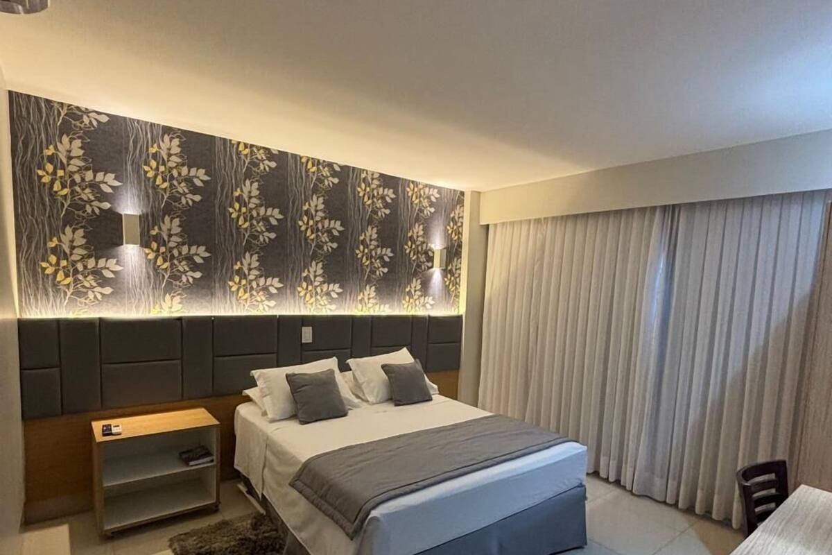 Un dormitorio moderno con una cama grande, una mesita de noche, una lámpara de pared y un ventanal con cortinas.