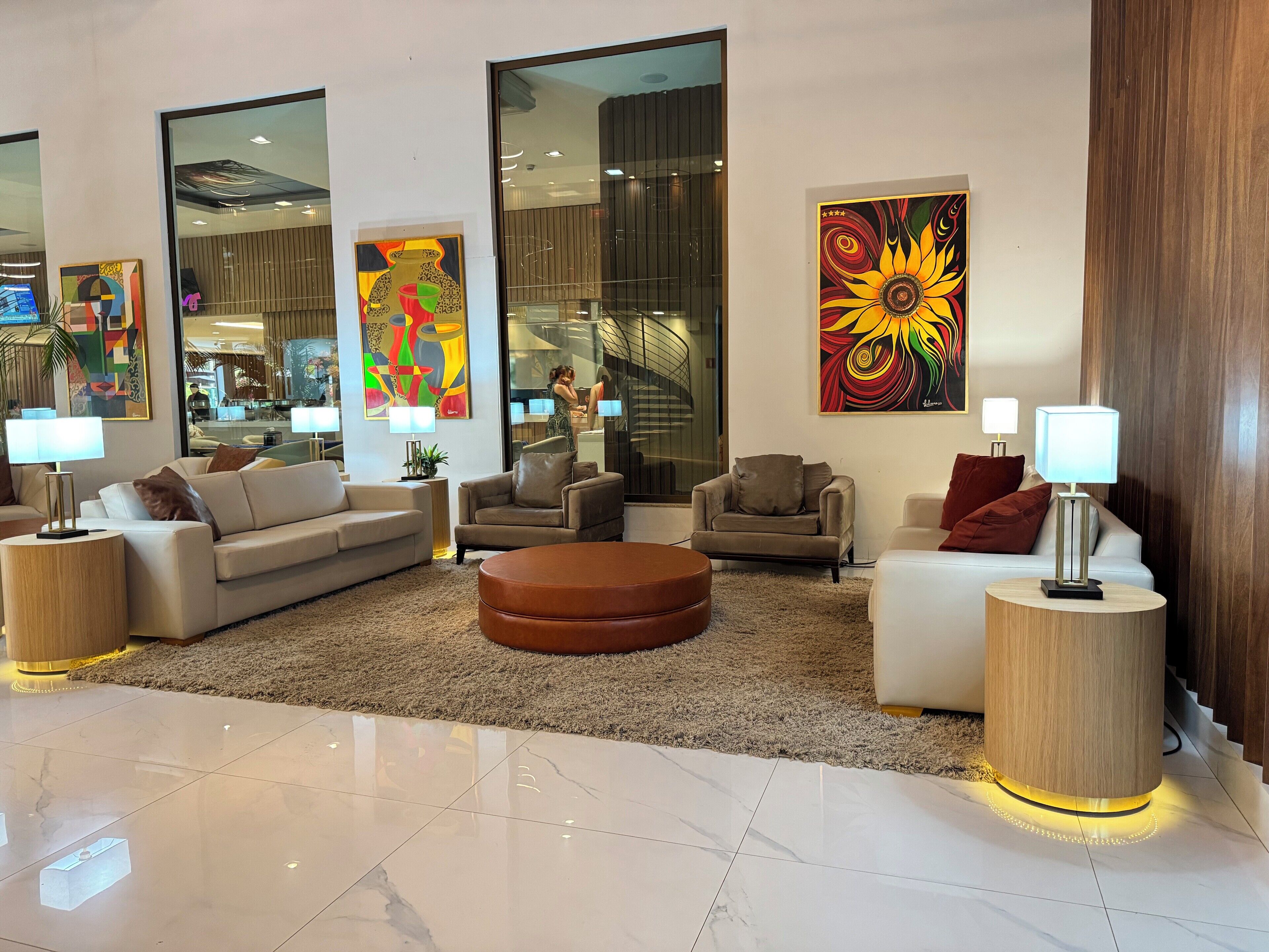Uma sala de estar moderna com um sofá, poltronas e uma mesa de centro. Há quadros abstratos nas paredes e uma grande janela com vista para o interior.