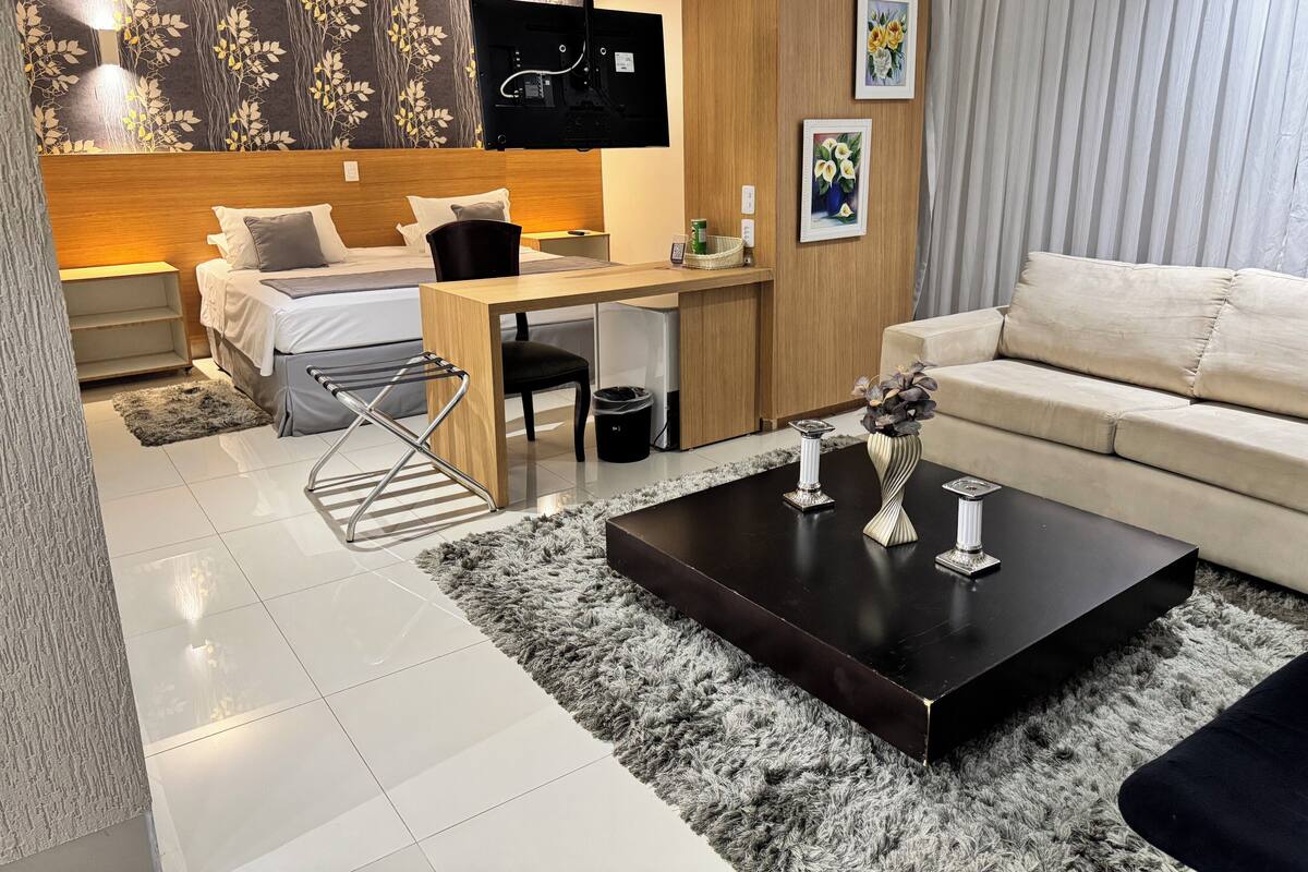 Una habitación de hotel moderna con cama, escritorio y sofá. Hay un televisor montado en la pared, una mesita con un florero y una ventana con cortinas.