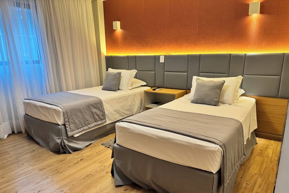Dos camas individuales con ropa de cama gris, una mesita de noche de madera y un aplique fijado a la pared.