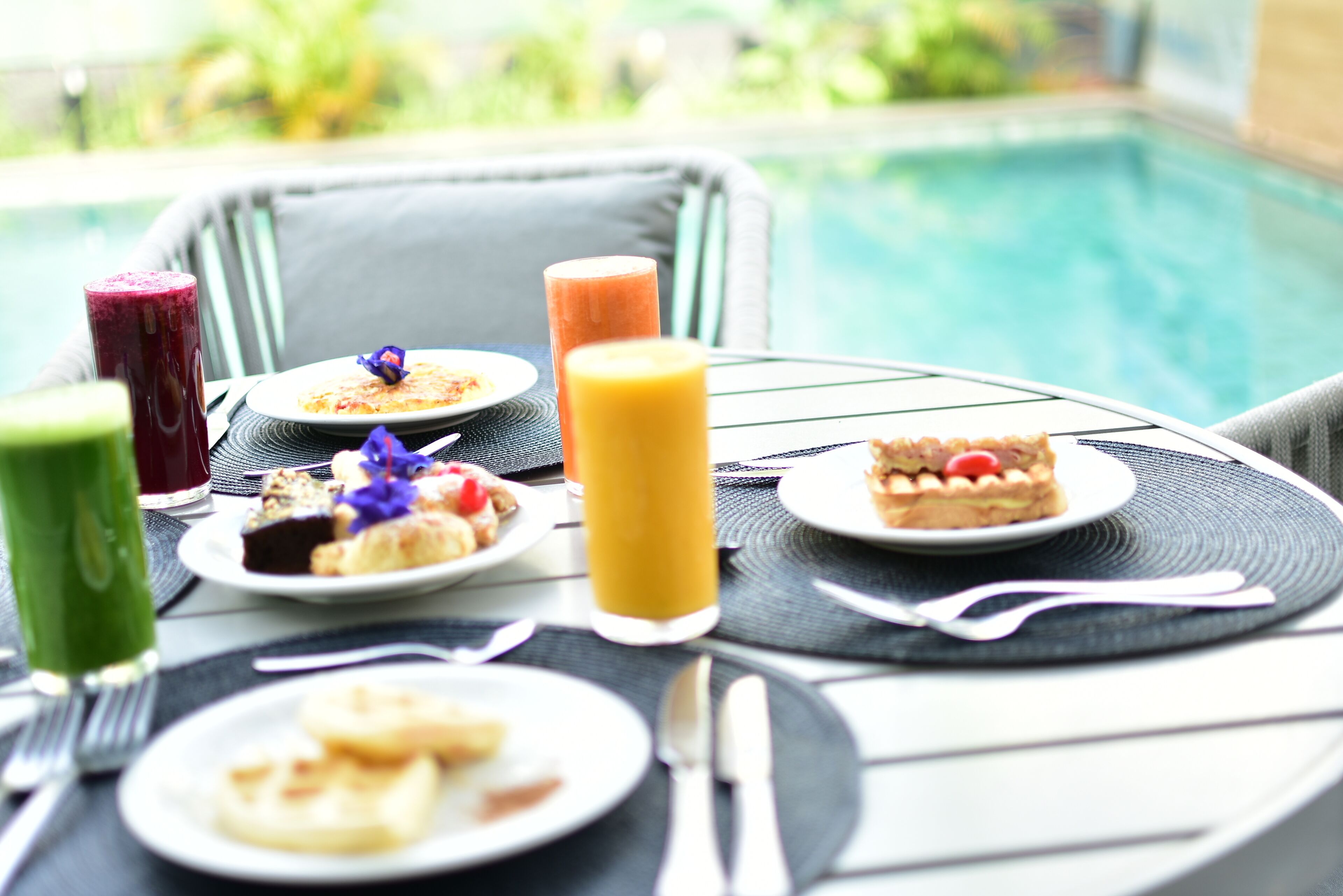 Uma mesa de jantar com pratos de comida e bebidas, piscina ao fundo, e uma cadeira com almofada.