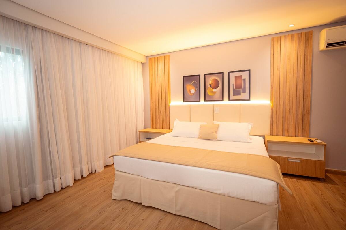 Una habitación de hotel moderna con una cama grande, cabecera de madera, cortinas blancas y tres cuadros en la pared.