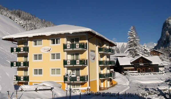 Un hotel de varios pisos con balcones, una pista de esquí al fondo y árboles cubiertos de nieve.