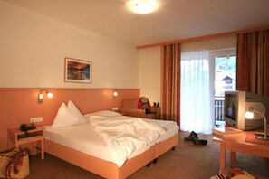 Una habitación de hotel con cama, escritorio con televisor, silla, maleta y ventana con cortinas.
