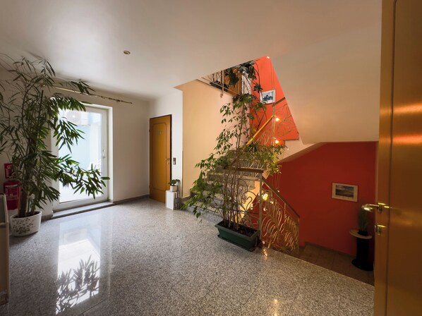 A spacious hallway with a polished marble floor, a wooden door, and a staircase with a red wall and decorative plants.