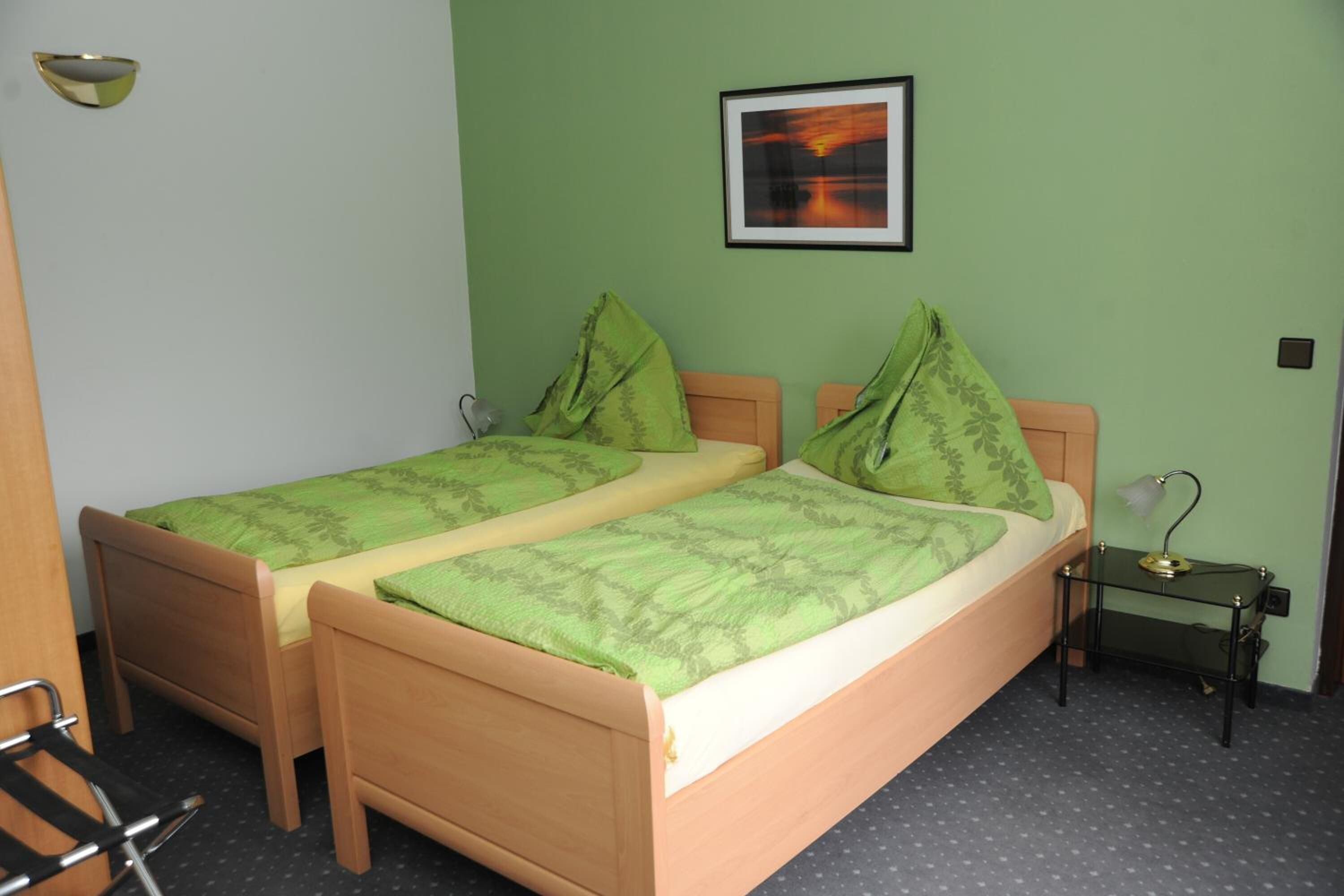 Una habitación con dos camas de madera, ropa de cama verde, un cuadro en la pared y una pequeña mesa con una lámpara.