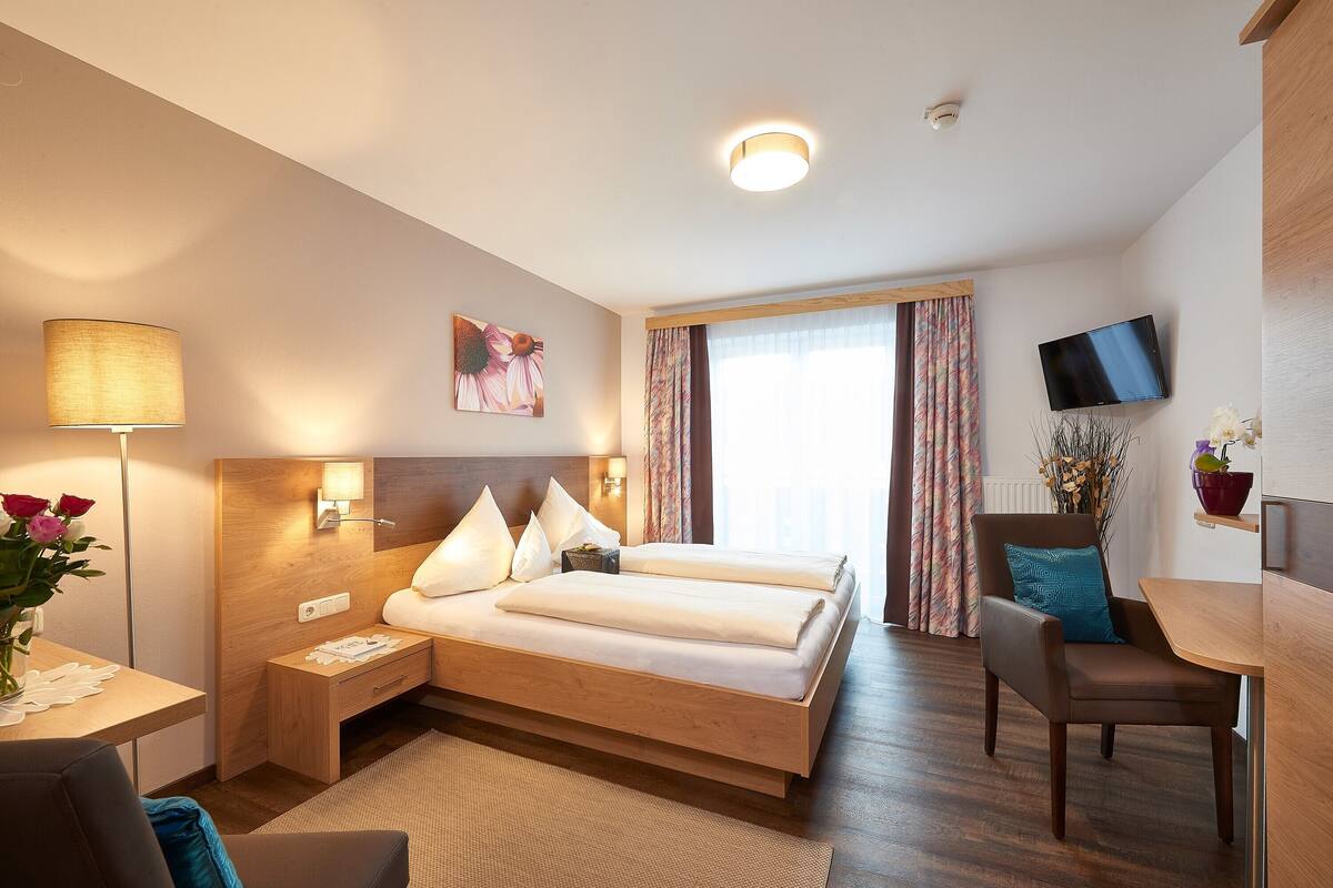 Een hotelkamer met een bed, nachtkastje, lamp, televisie en een klein tafeltje met stoel.