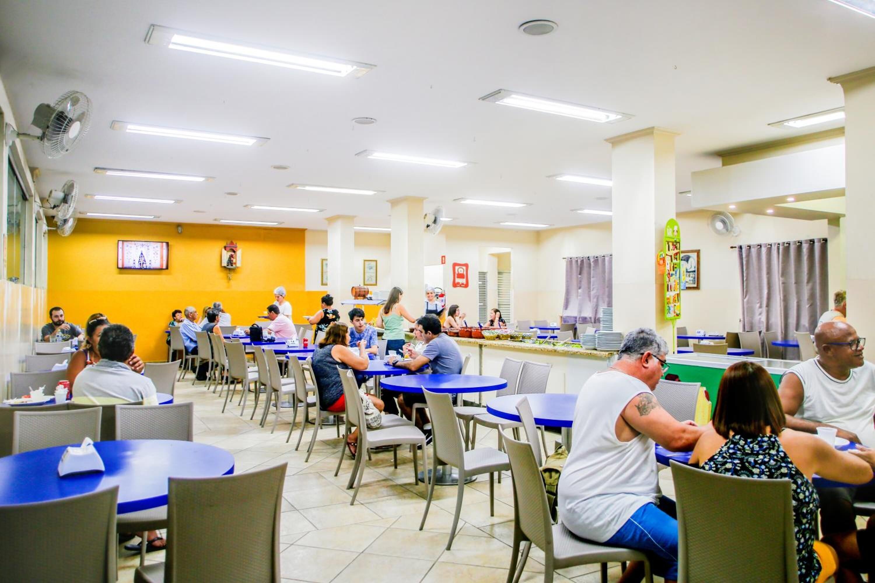 A cafeteria with multiple tables and chairs, a television mounted on the wall, and a counter with food items.