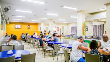 A cafeteria with multiple tables and chairs, a television mounted on the wall, and a counter with food items.