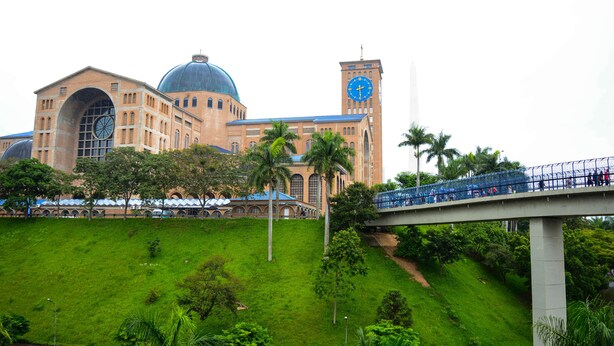 A large building with a clock tower and a dome, surrounded by greenery and a pedestrian bridge.