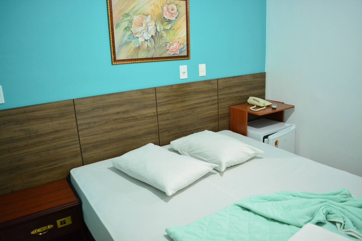 A hotel room with a bed, pillows, a nightstand, a telephone, and a painting on the wall.