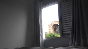 A room with a window, a bed, and a view of a building with a clock.