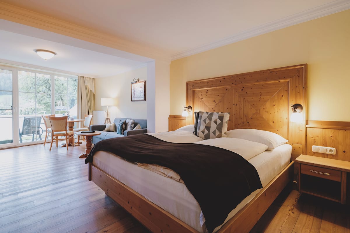 A bedroom with a large bed, wooden headboard, bedside tables, a nightstand, a chair, a small table, and a window with curtains.
