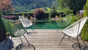 Een houten terras met witte, metalen stoelen met uitzicht op een rustig meer, omringd door weelderige begroeiing en bergen.