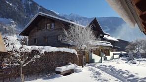 A wooden chalet with a sloped roof, surrounded by snow-covered trees and mountains in the background.