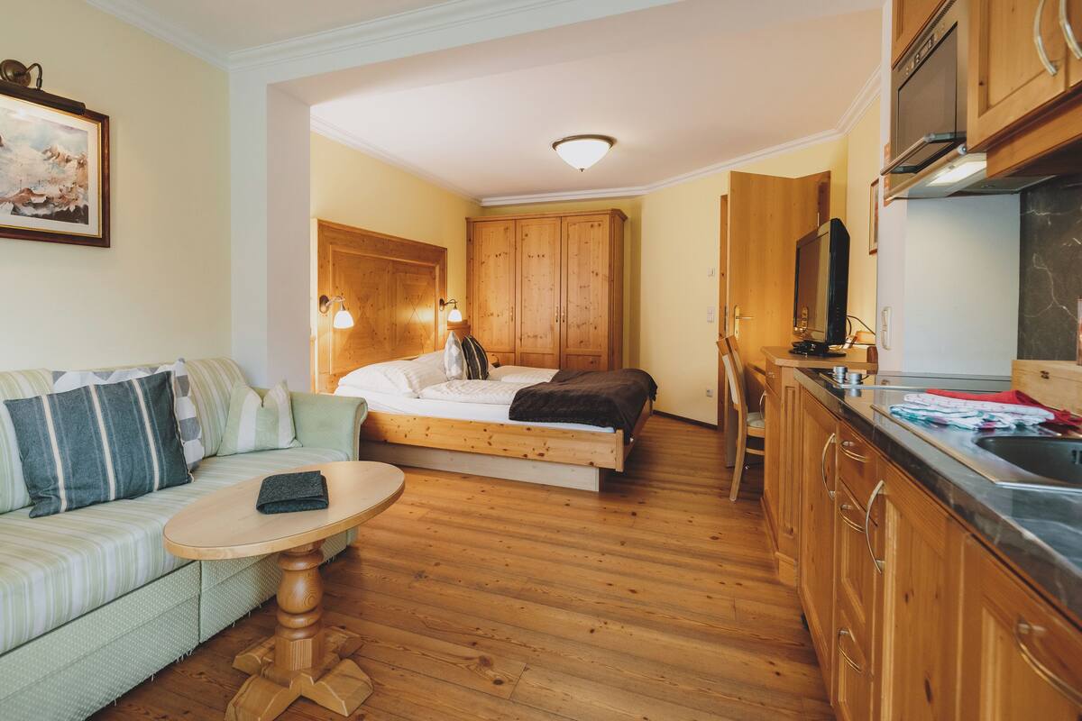 A compact room with a bed, a sofa, a small table, wooden cabinets, and a kitchenette.