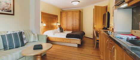 A compact room with a bed, a sofa, a small table, wooden cabinets, and a kitchenette.
