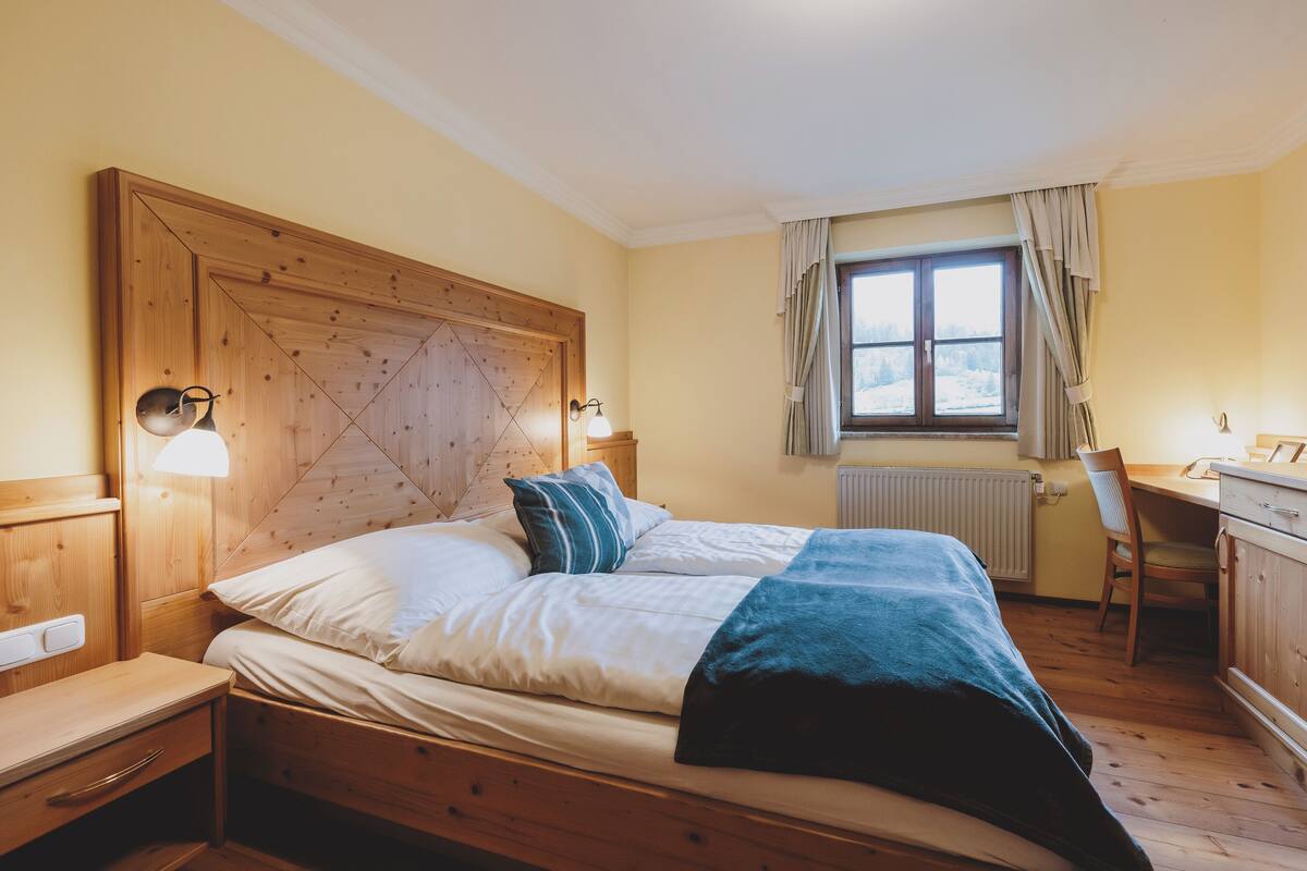 A bedroom with a wooden headboard, a bed with white and blue bedding, a desk with a chair, and a window with curtains.