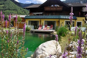 Hotel frente a un lago con muelle de madera, asientos al aire libre y montañas al fondo.