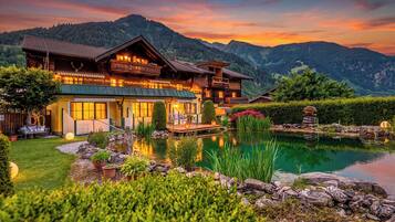 Un chalet de madera grande con un estanque, rodeado de montañas y vegetación exuberante.