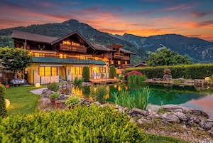 A large wooden chalet with a pond, surrounded by mountains and lush greenery.