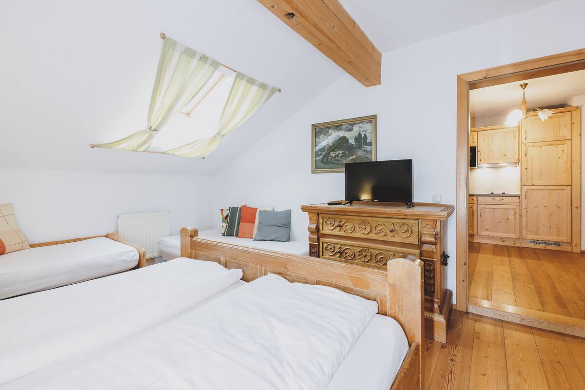 A bedroom with two beds, a wooden dresser, a painting, and a view into a kitchen.