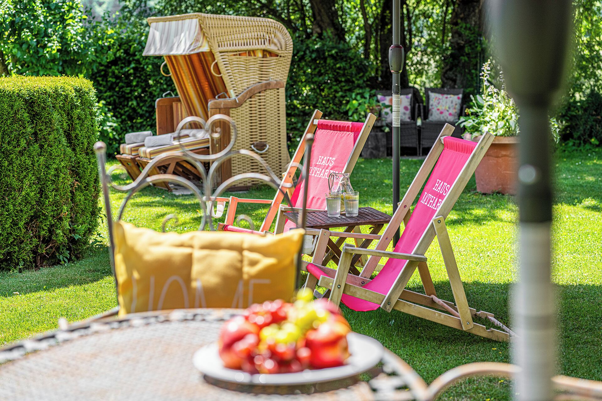 A garden setting with outdoor furniture, including a wicker chair, a metal chair, and a table with a bowl of fruit.