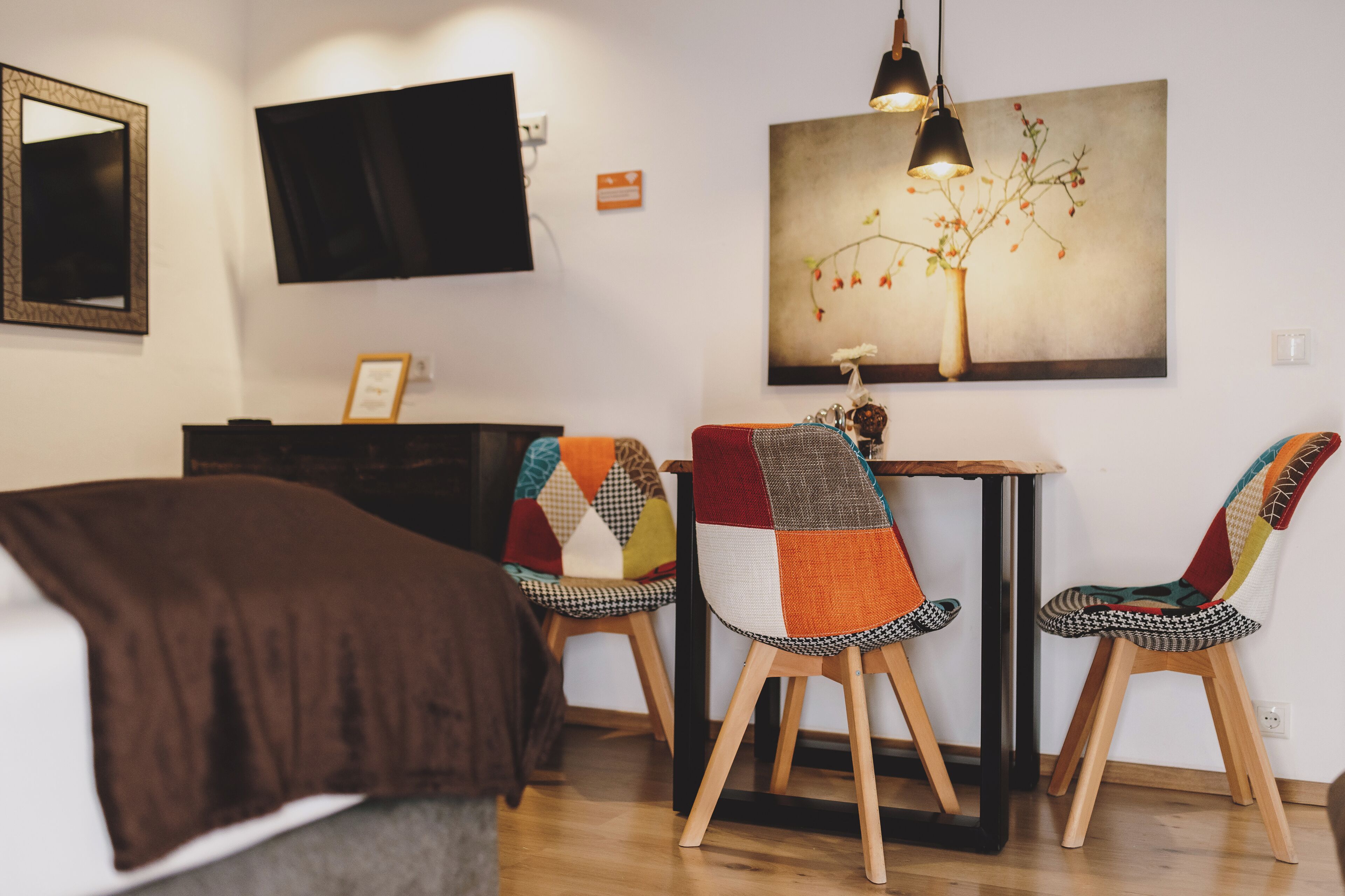 A modern room with a bed, a desk, two chairs with colorful upholstery, a painting of a tree, and a TV mounted on the wall.