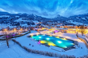 Paisaje nevado con una gran piscina, edificios iluminados y montañas al fondo.