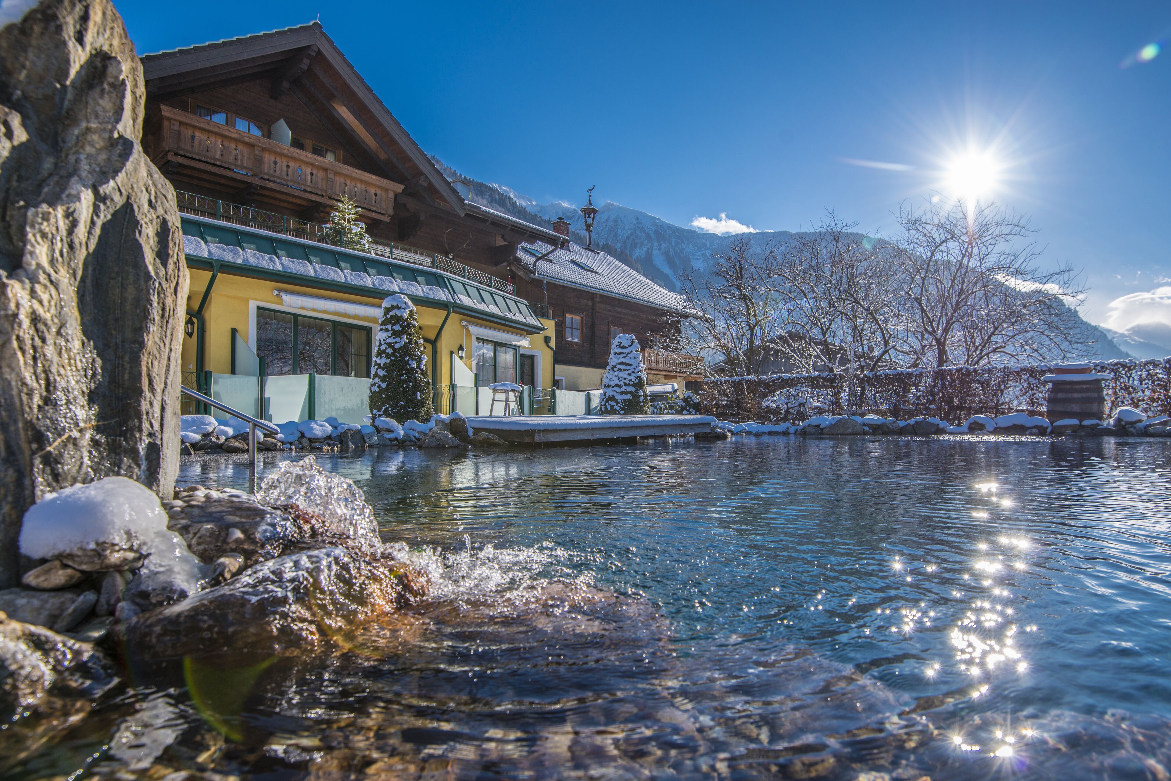 A wooden chalet with a balcony, a pool, and snow-covered mountains in the background.