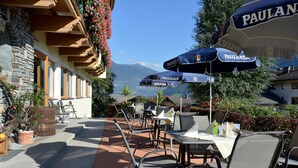 An outdoor patio with tables and chairs, umbrellas, and a stone building with wooden beams. - Hotel - Gasthof Jäger Heim GmbH (Schlitters)