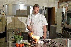 Un chef cocina a fuego vivo en una estufa en una cocina con electrodomésticos de acero inoxidable y un salpicadero de azulejos.