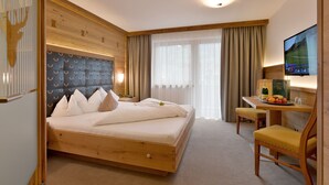 A hotel room with a bed, a desk with a chair, a television, and a window with curtains. - Hotel - Gasthof Jäger Heim GmbH (Schlitters)