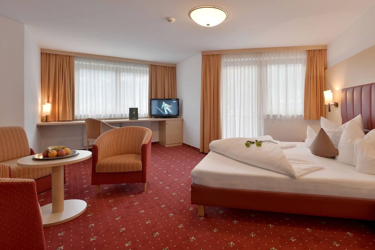 Een hotelkamer met een bed, een bureau met televisie, een stoel, een klein tafeltje met fruit en een nachtkastje met een fles en een glas.