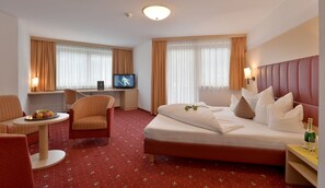 Une chambre d’hôtel avec un lit, un bureau avec une télévision, une chaise, une petite table avec des fruits et une table de chevet avec une bouteille et un verre.