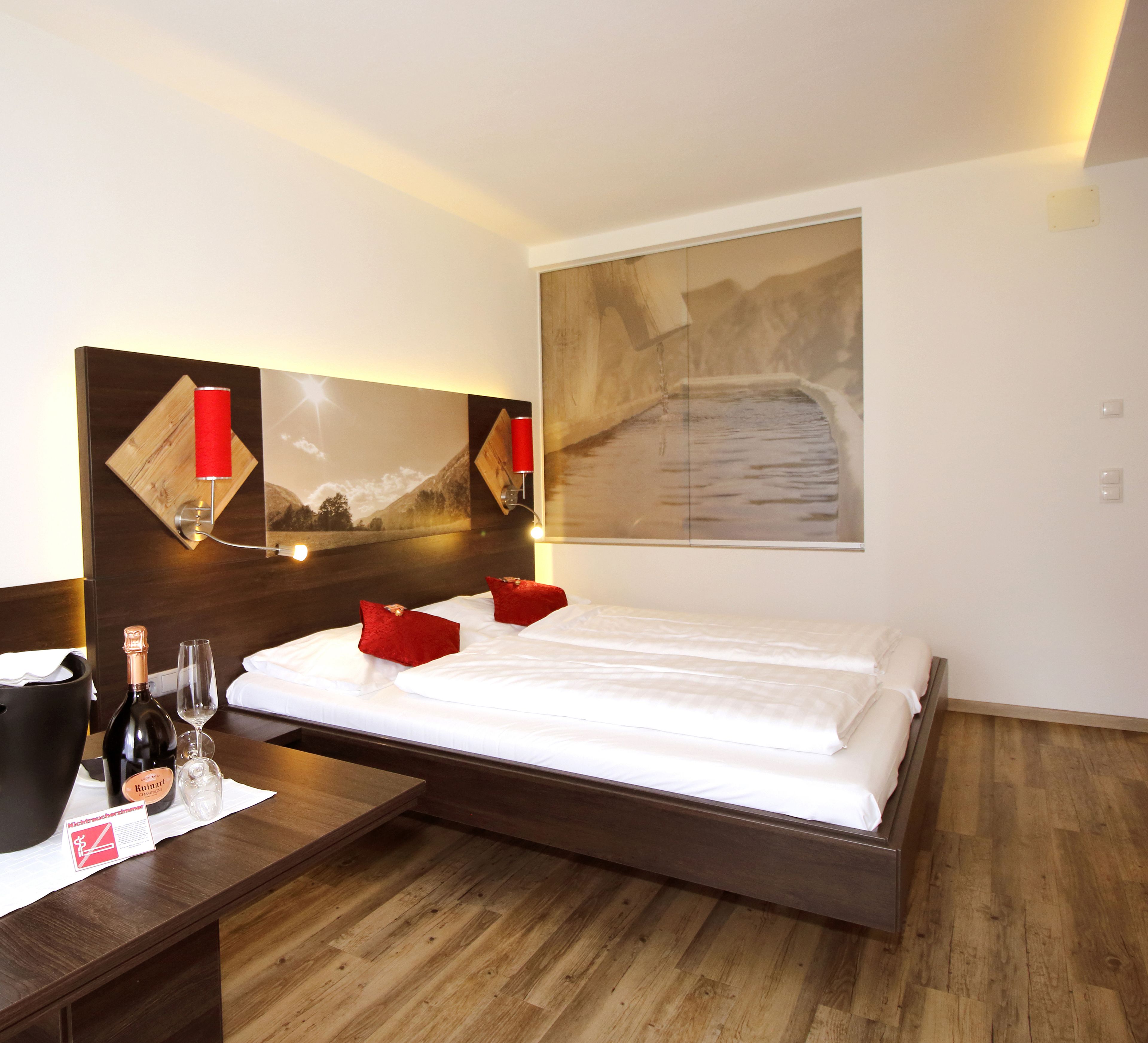 A hotel room with a wooden bed, a large painting on the wall, and a bedside table with a bottle of wine and glasses.