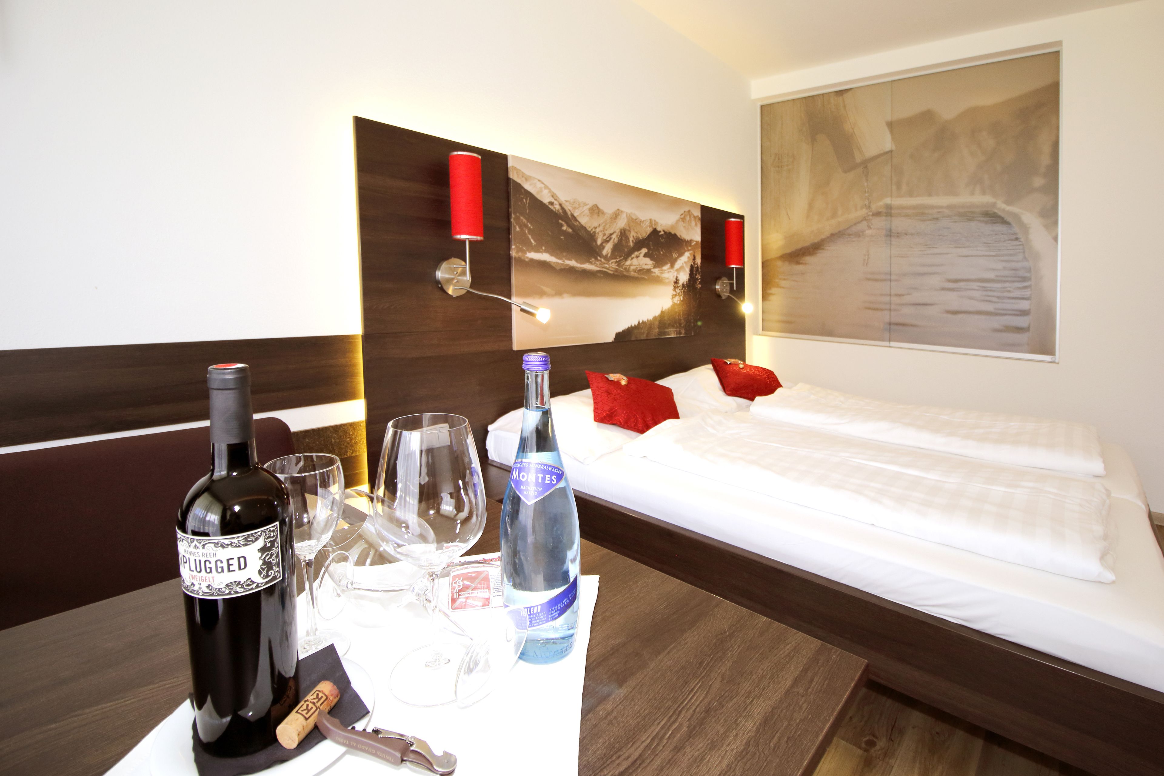 A hotel room with a bed, a nightstand with a wine bottle and glasses, a painting on the wall, and a view of mountains.