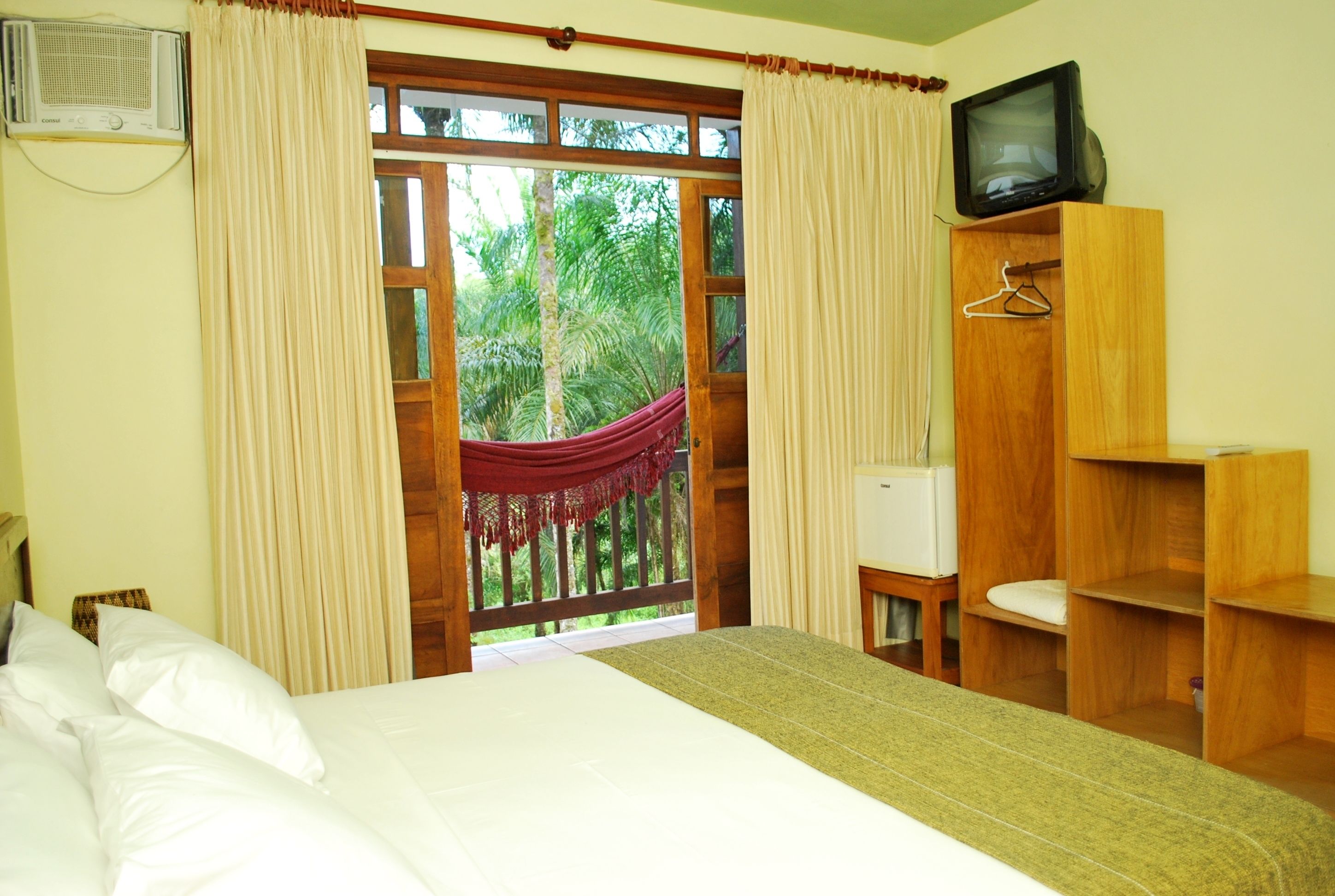 A bedroom with a bed, a television on a wooden cabinet, a hanging chair, and a view of greenery outside.