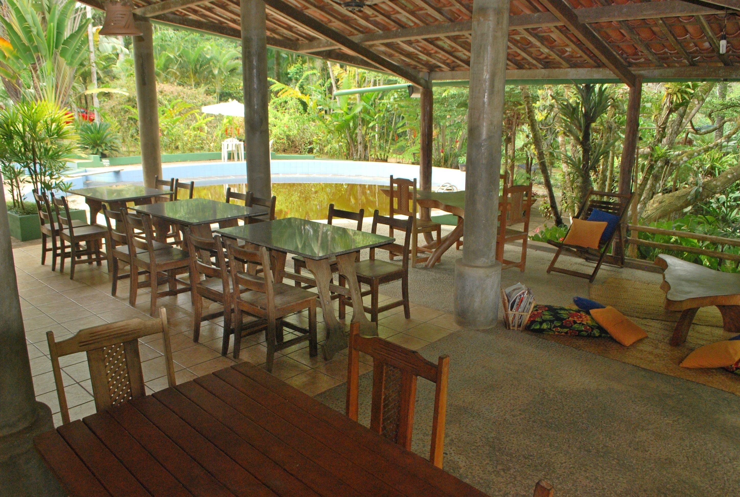 Un patio al aire libre con mesas y sillas de madera, una piscina y abundante vegetación.