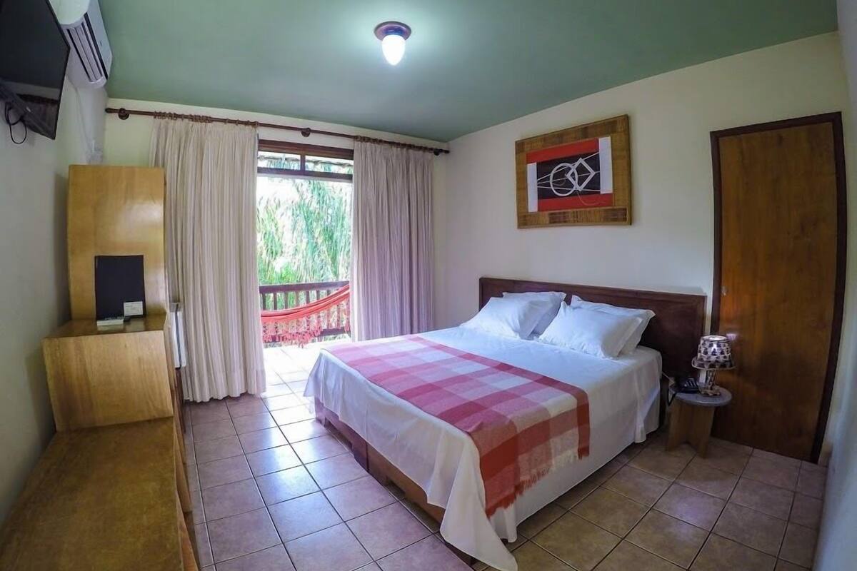 Un dormitorio con cama, mesita de noche, lámpara, aparador de madera, un cuadro y vistas al exterior con vegetación.