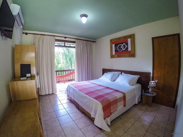 Un dormitorio con cama, mesita de noche, lámpara, aparador de madera, un cuadro y vistas al exterior con vegetación.