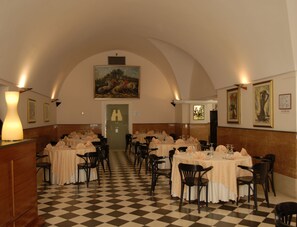 Un espace repas doté d’un sol à carreaux, de tables recouvertes de nappes blanches et de chaises noires, ainsi que d’un grand tableau accroché au mur.